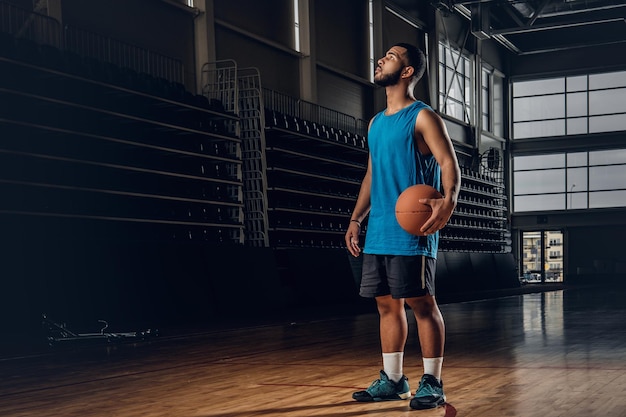 Portrait of Black basketball player holds a ball over a hoop in a basketball hall.