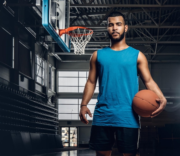 Portrait of Black basketball player holds a ball over a hoop in a basketball hall.