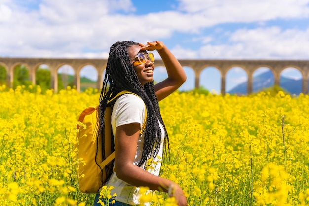 Portrait of a black ethnic girl with braids looking at the sun sunglasses traveler in a field of yellow flowers