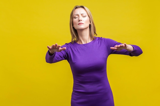 Portrait of blind disoriented young woman in elegant tight purple dress walking with closed eyes reaching out hands to find lost way vision loss indoor studio shot isolated on yellow background