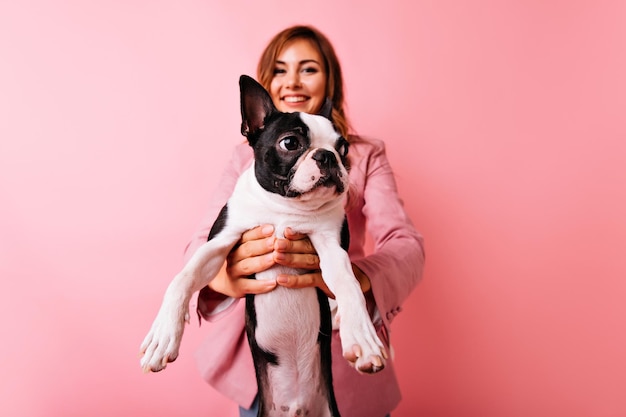 Portrait of blissful white girl with french bulldog on foreground Indoor photo of goodlooking female model posing with cute little dog