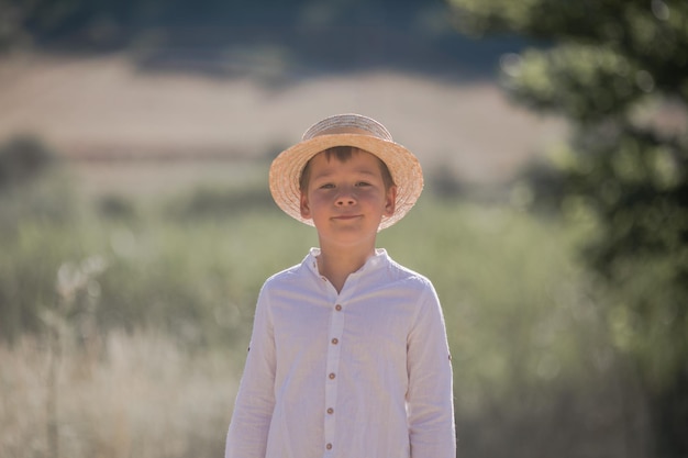 Portrait of the blode hair boy in straw hat with blurred summer greenery background