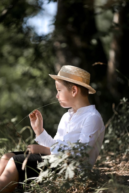 Portrait of the blode hair boy in straw hat with blurred summer greenery background