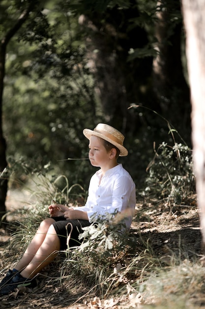 Portrait of the blode hair boy in straw hat with blurred summer greenery background