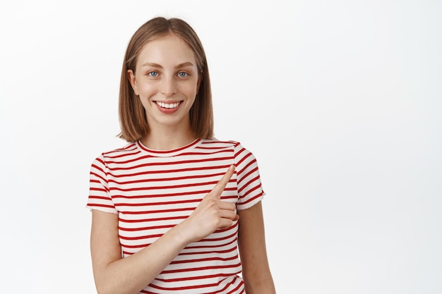 Portrait of blond happy woman smiling, pointing finger at upper right corner, showing sale discount promo, product placement, standing against white wall