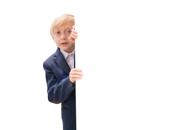 Portrait of blond little boy with white blank isolated on white background Boy with glasses and school uniform peeking out from behind board