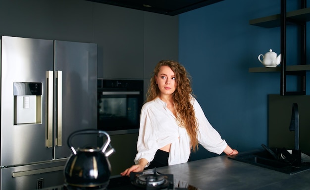 Portrait of blonde caucasian woman standing and smiling in her kitchen leaning on the table young