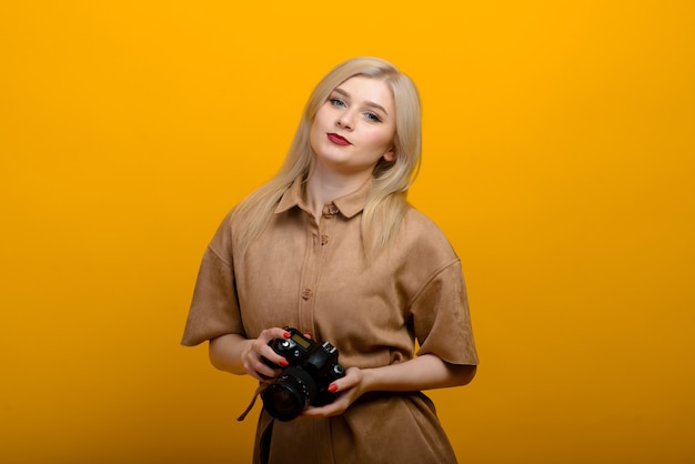 Portrait of a blonde girl with a camera in hand on a yellow background. Isolated studio.