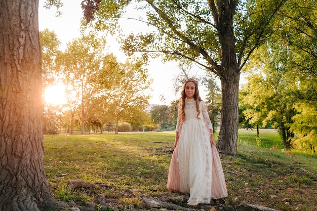 Portrait of a blonde girl with curly hair dressed in communion dress walking through some trees