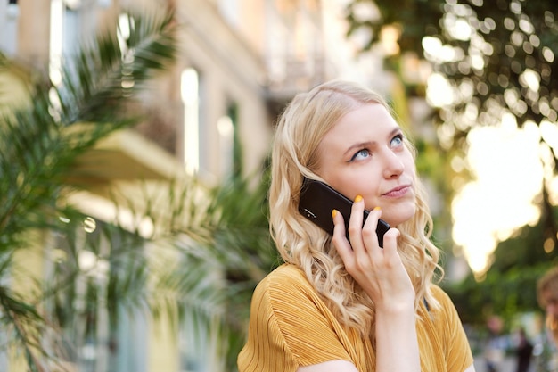 Portrait of bored blond girl thoughtfully looking away while talking on cellphone on city street