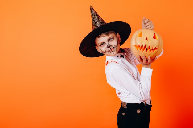 Portrait boy in Devil masquerade makeup and hat  showing a pumpkin. Halloween 