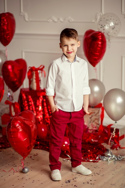 portrait of a boy in a White Shirt and red trousers on the background of the festive decor of red balls in the widget of hearts and gifts. holiday birthday or valentines day