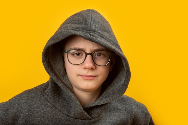 Portrait of boy with glasses wears hoodie and hood on yellow background Shy unsociable teenager