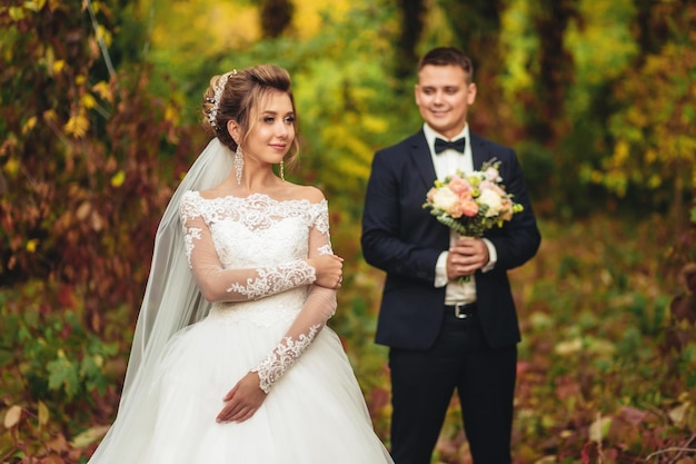 Portrait of a bride in an autumn park The groom looks at the bride in the background with tenderness