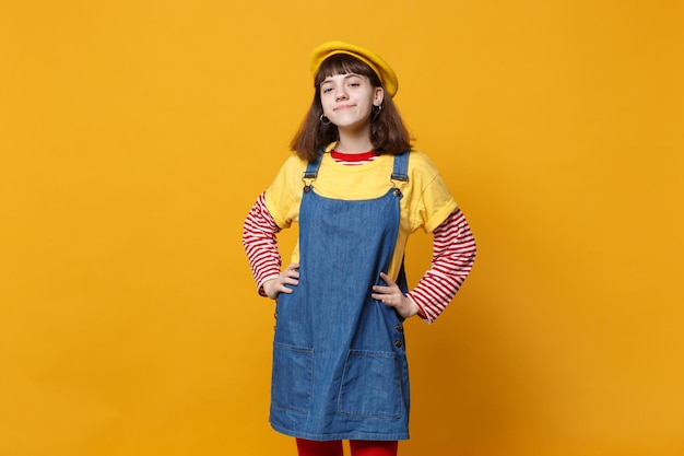 Portrait of bright girl teenager in french beret, denim sundress standing with arms akimbo on waist isolated on yellow wall background. People sincere emotions, lifestyle concept. Mock up copy space.