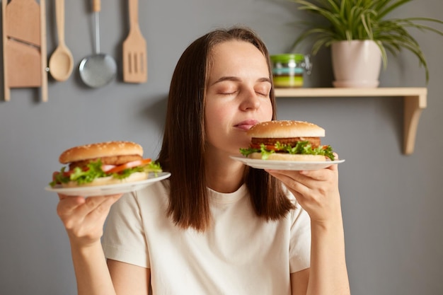 Portrait of brown haired Caucasian woman holding two delicious fresh hamburgers smelling aromat of fast food with closed eyes and satisfied facial expression