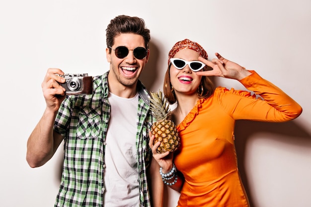 Portrait of brunet man taking picture on retro camera and woman in orange dress and headband Girl in glasses holds pineapple and shows peace sign