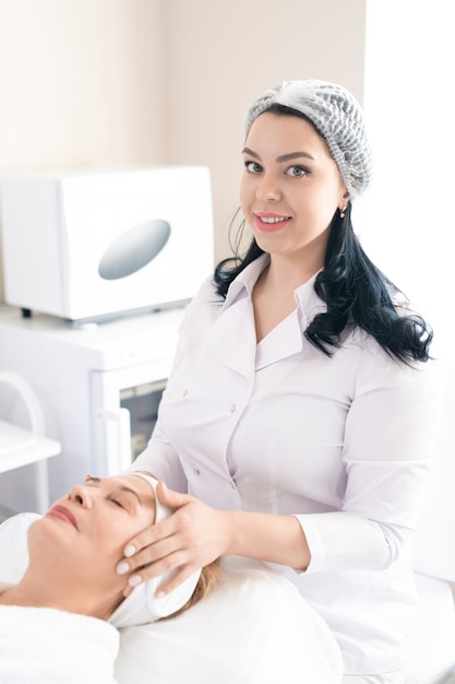 Portrait of brunette beautician in white coat sitting at procedure table and giving face sculpting massage to mature woman