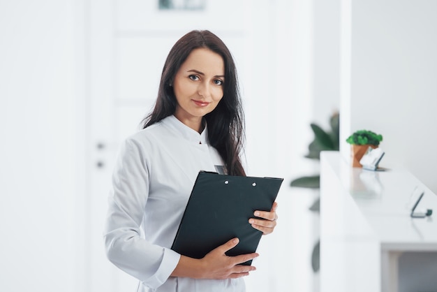 Portrait of brunette doctor with notepad in hands that stands indoors at clinic.