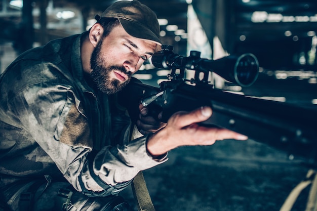 Portrait of brunette is taking aim. He is looking through lens. Guy is holding rifle with right hand and keeping his left one on trigger. Man is ready to shoot.