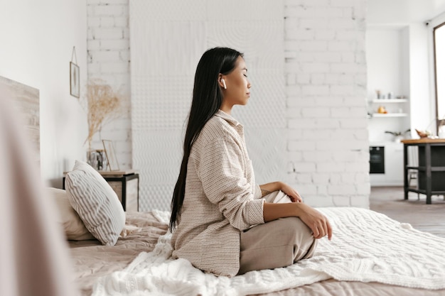 Portrait of brunette longhaired Asian woman in beige jacket and pants meditating sitting on soft bed and listening relaxing music in headphones