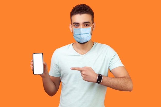 Portrait of brunette man with medical mask in white t-shirt pointing at cellphone and smiling at camera, recommending gadget or mobile application. indoor studio shot isolated on orange background