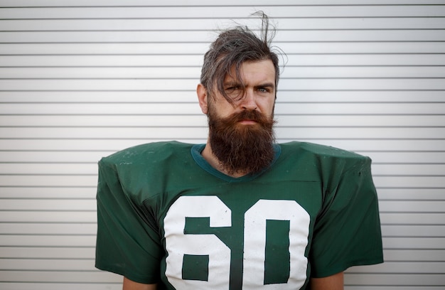 Portrait of brutal bearded man in green uniform for playing american football against white wall