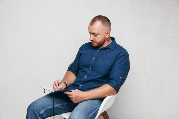 Portrait of a brutal man sitting on a chair against a white wall with a laptop and notepada business man