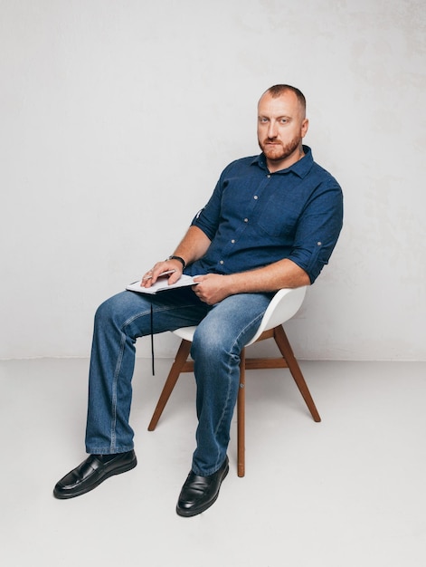 Portrait of a brutal man sitting on a chair against a white wall with a laptop and notepada business man