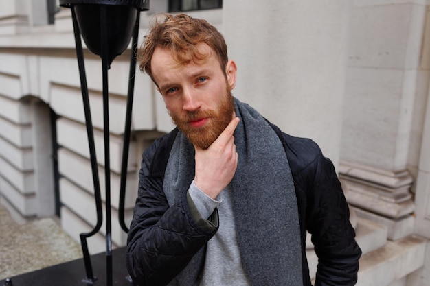 Portrait of a brutal redhaired man in a jacket and scarf on the street near a beautiful building