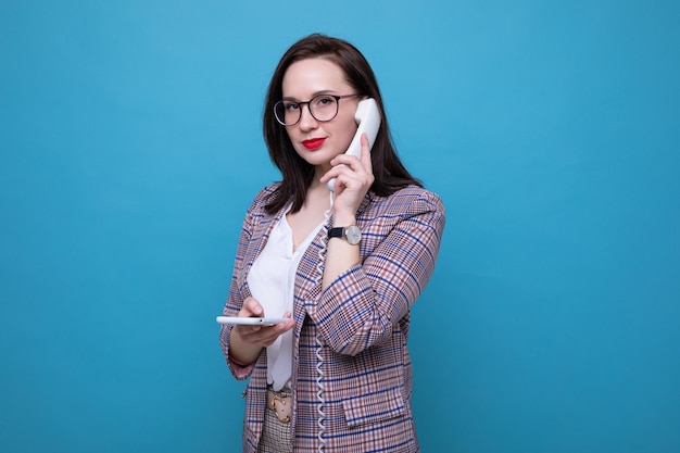 Portrait of a business woman who is talking on an office phone and holding a smartphone in her hand