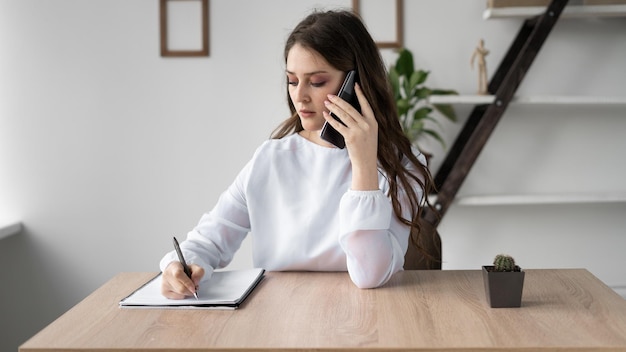 Portrait of a business woman working from home sitting at a desk talking or talking on the phone with a client and making notes in a notebook Mobile network consulting office work