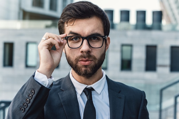 Portrait of a businessman is touching eyeglasses in a classic suit against an office building. 