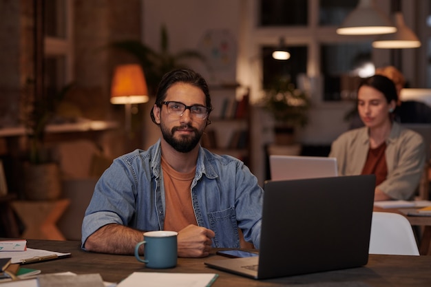 Portrait of businessman looking at front while sitting at his workplace with laptop and working at office with his colleagues in the wall