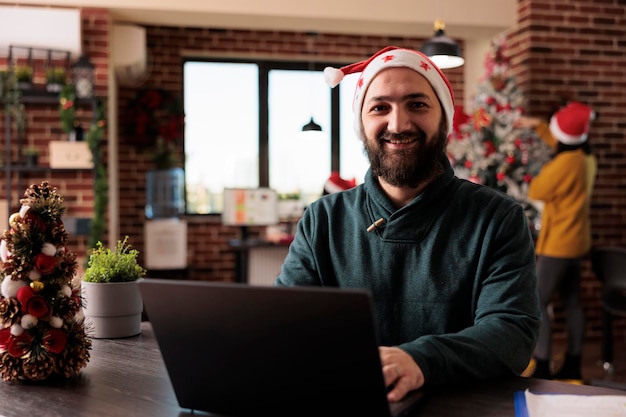 Portrait of businessman working on company laptop and sitting in office decorated with christmas tree and lights, festive ornaments. Smiling worker wearing santa hat during seasonal xmas time.