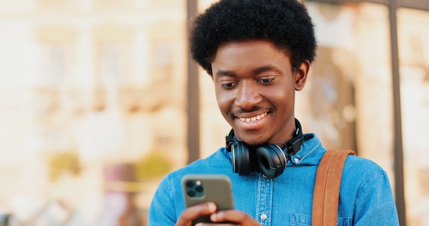 Portrait of calm brunette African American man wearing casual clothes looking at the smartphone screen while standing on green street spring day and showing his teeth at the wide smile