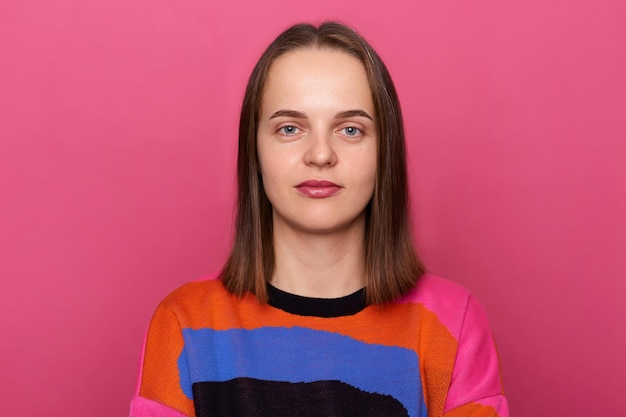 Portrait of calm cute attentive young adult Caucasian woman with brown hair wearing colorful jumper posing isolated over pink background looking at camera