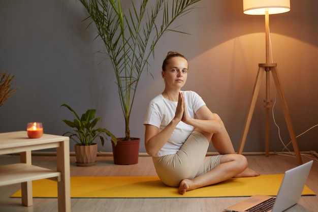 Portrait of calm relaxed woman with hair bun in t shirt practicing yoga using laptop for working with her couch online sitting on floor holding hands in namaste gesture and meditating relaxing