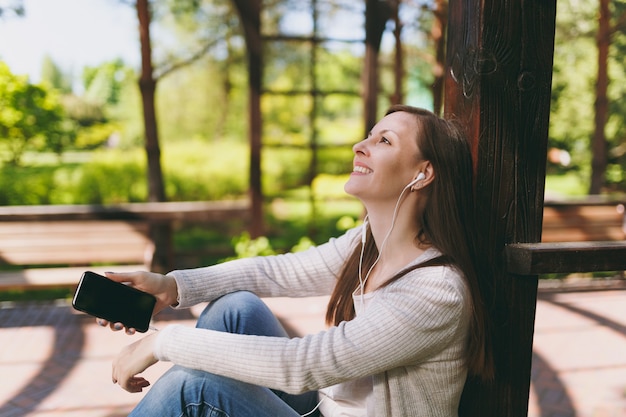 Portrait of calm woman in casual clothes. Female sitting in park in street outdoors on nature, listening music in mobile phone with blank empty screen to copy space in earphones. Lifestyle concept.