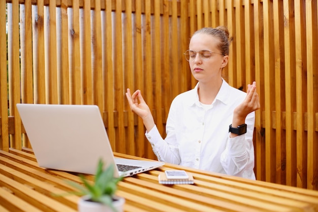 Portrait of calm young adult dark haired woman with hair bun wearing white shirt sitting and working on laptop trying to calm down meditating practicing yoga during break