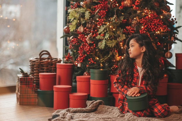 Portrait of candid asian smiling little girl in red plaid pajama sitting with presents at Xmas home