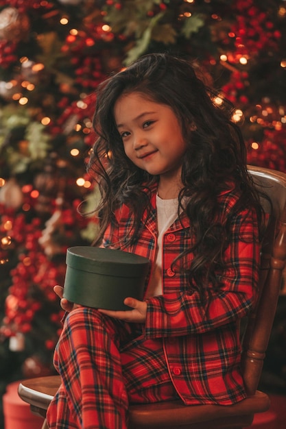 Portrait of candid asian smiling little girl in red plaid pajama sitting with presents at Xmas home