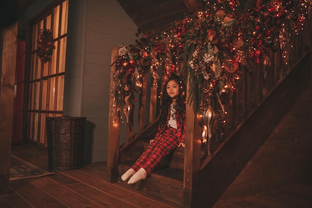 Portrait of candid asian smiling little girl in red plaid pajama sitting on wooden steps at Xmas