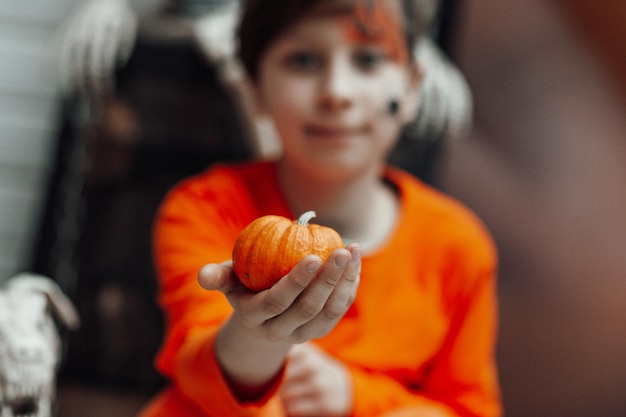 Portrait of candid authentic teen boy with face painted holds pumpkin at Halloween decorated party