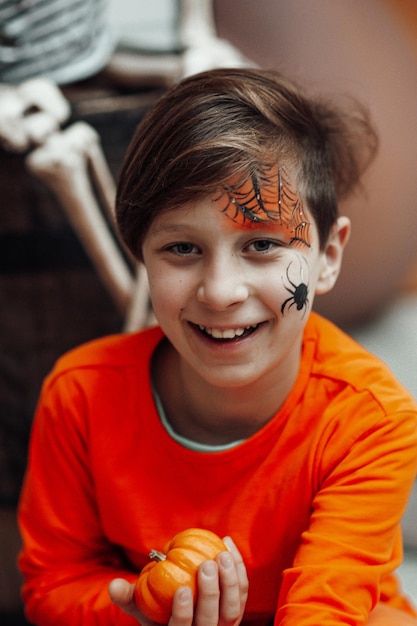 Portrait of candid authentic teen boy with face painted holds pumpkin at Halloween decorated party