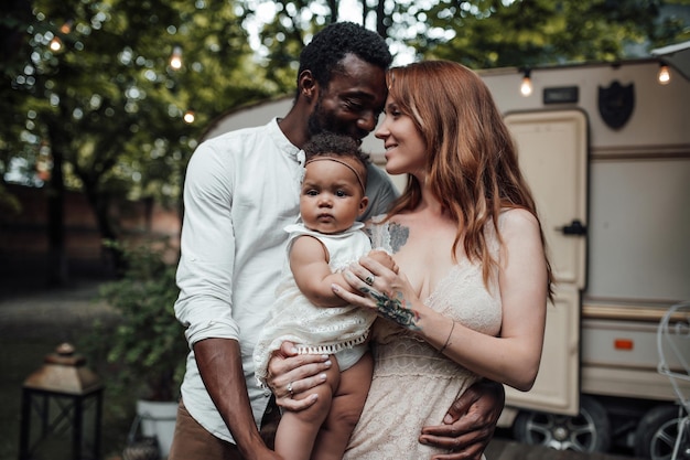 Portrait of candid friendly happy interracial family with swarthy baby outdoor near travel trailer