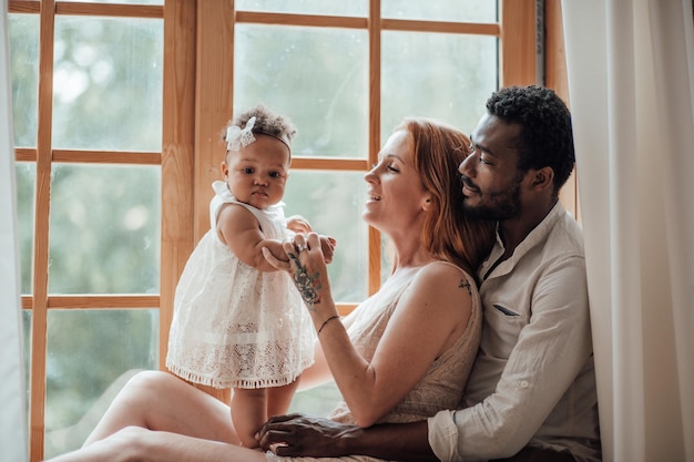 Portrait of candid friendly happy interracial family with swarthy baby together near window