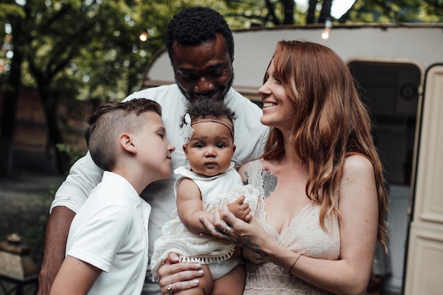 Portrait of candid friendly happy interracial family with two kids outdoor near retro travel trailer