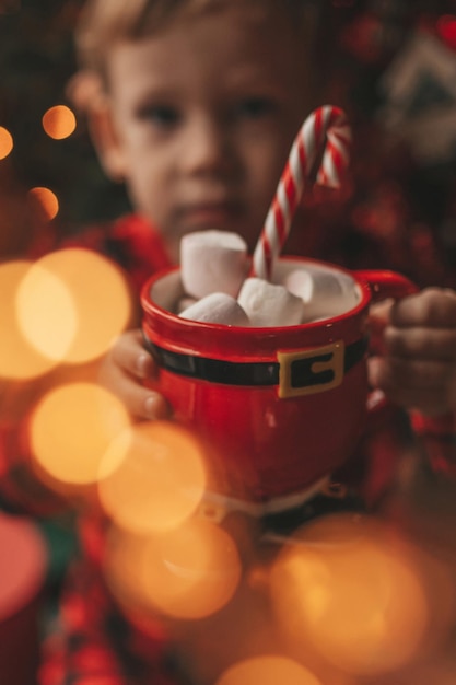 Portrait candid happy child in red plaid pajama hold Xmas mug with marshmallows and candy cane