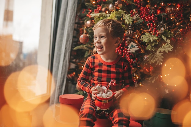 Portrait candid happy child in red plaid pajama hold Xmas mug with marshmallows and candy cane